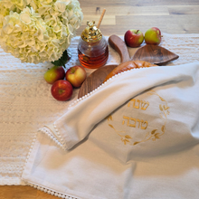 White, woven cotton-blend challah cover with petite pom-pom fringe and gold embroidered Shanah Tova text in Hebrew encircled by a leafy gold embroidered wreath design. Shown on an apple-shaped wooden challah board alongside a glass honey server and several ripe apples.