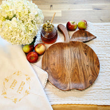 White, woven cotton-blend challah cover with petite pom-pom fringe and gold embroidered Shanah Tova text in Hebrew encircled by a leafy gold embroidered wreath design. Shown next to an apple-shaped wooden challah board alongside a glass honey server and several ripe apples.