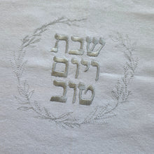 Silver embroidery spells out Shabbat v’Yom Tov in Hebrew lettering encircled by a leafy wreath in the same silver color.