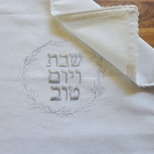 Closeup view of a woven Challah Cover with picot edging featuring center embroidery. Silver embroidery spells out Shabbat v’Yom Tov in Hebrew lettering encircled by a leafy wreath in the same silver color.