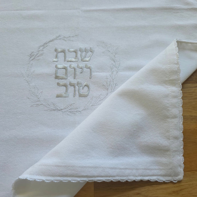 A white, woven Challah Cover with picot edging featuring center embroidery. Silver embroidery spells out Shabbat v’Yom Tov in Hebrew lettering encircled by a leafy wreath in the same silver color.