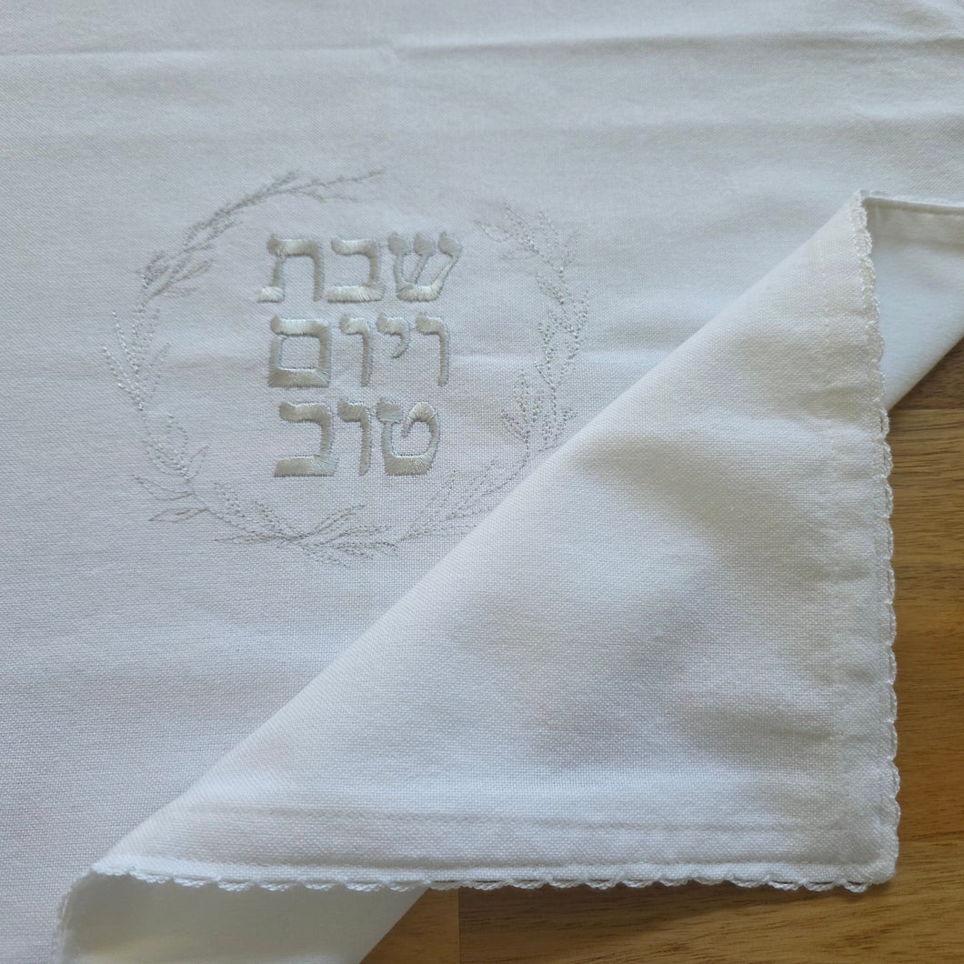 A white, woven Challah Cover with picot edging featuring center embroidery. Silver embroidery spells out Shabbat v’Yom Tov in Hebrew lettering encircled by a leafy wreath in the same silver color.
