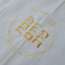 A zoomed in view of a white, woven Challah Cover with picot edging. Center embroidery spells out Shabbat v’Yom Tov in gold Hebrew lettering encircled by a leafy wreath in the same gold color.
