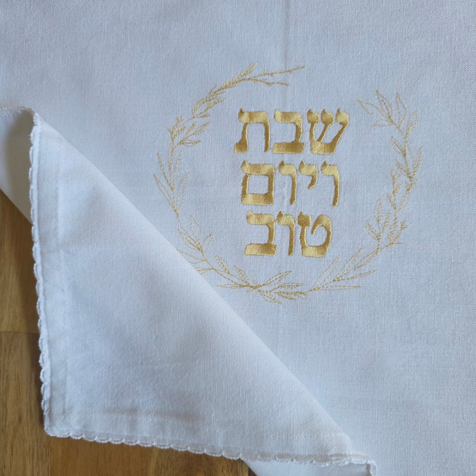 A white, woven Challah Cover with picot edging features center embroidery. Gold embroidery spells out Shabbat v’Yom Tov in Hebrew lettering encircled by a leafy wreath in the same gold color.