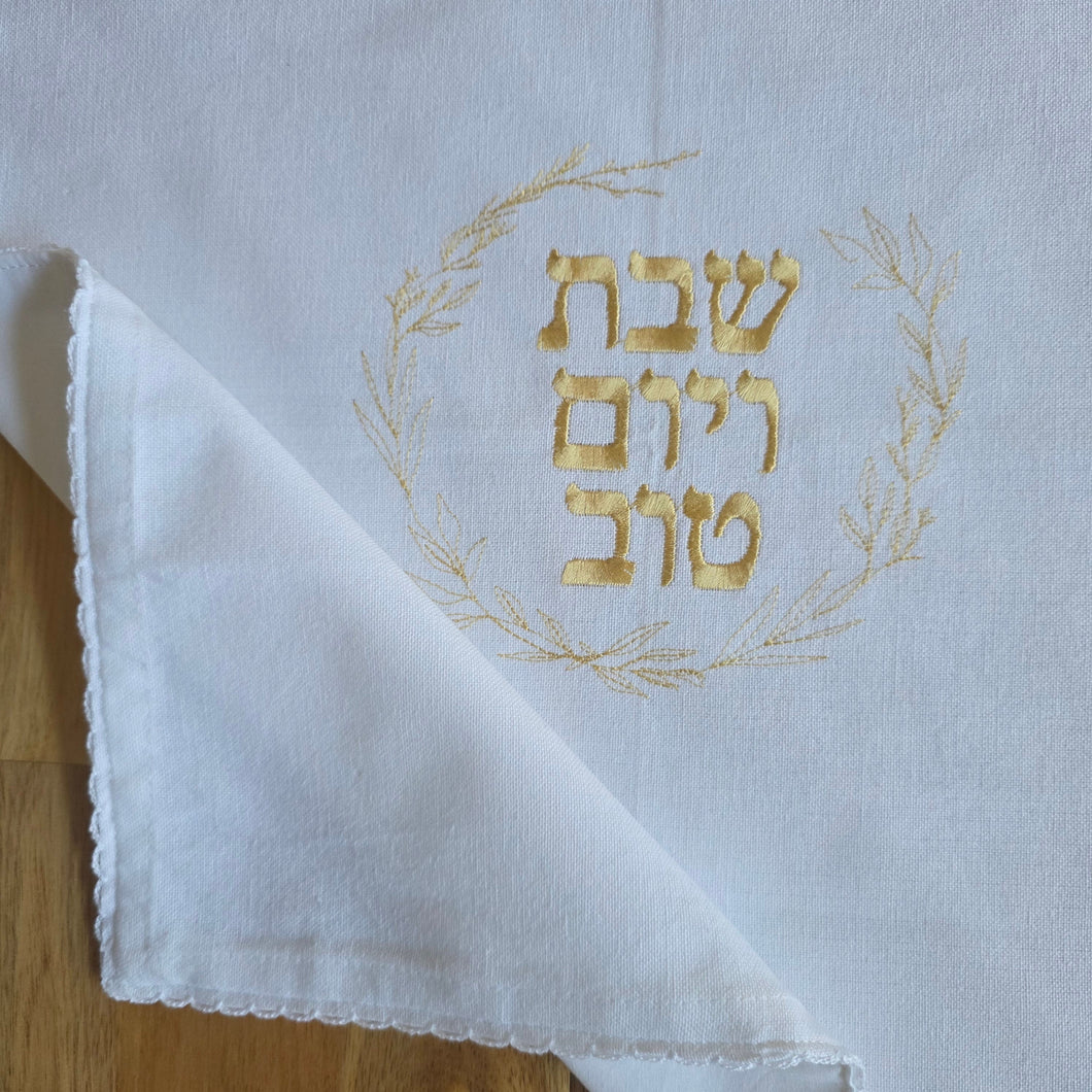 A white, woven Challah Cover with picot edging features center embroidery. Gold embroidery spells out Shabbat v’Yom Tov in Hebrew lettering encircled by a leafy wreath in the same gold color.