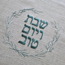 A woven natural challah cover with blue sage embroidery in a leafy wreath design and the words Shabbat v’Yom Tov in Hebrew. 