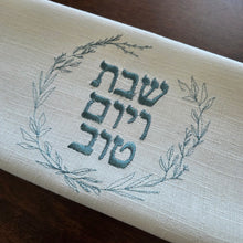 A woven cotton-blend challah cover featuring blue sage embroidered wreath with the message Shabbat v’Yom Tov in Hebrew lettering. Measures 18” x 18”.