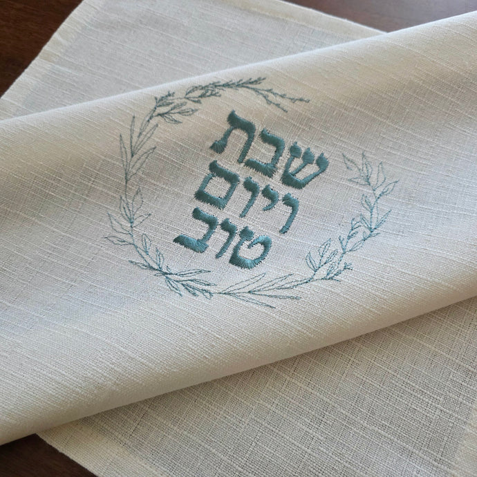A woven cotton-blend challah cover featuring blue sage embroidered wreath with the message Shabbat v’Yom Tov in Hebrew lettering. Measures 18” x 18”.