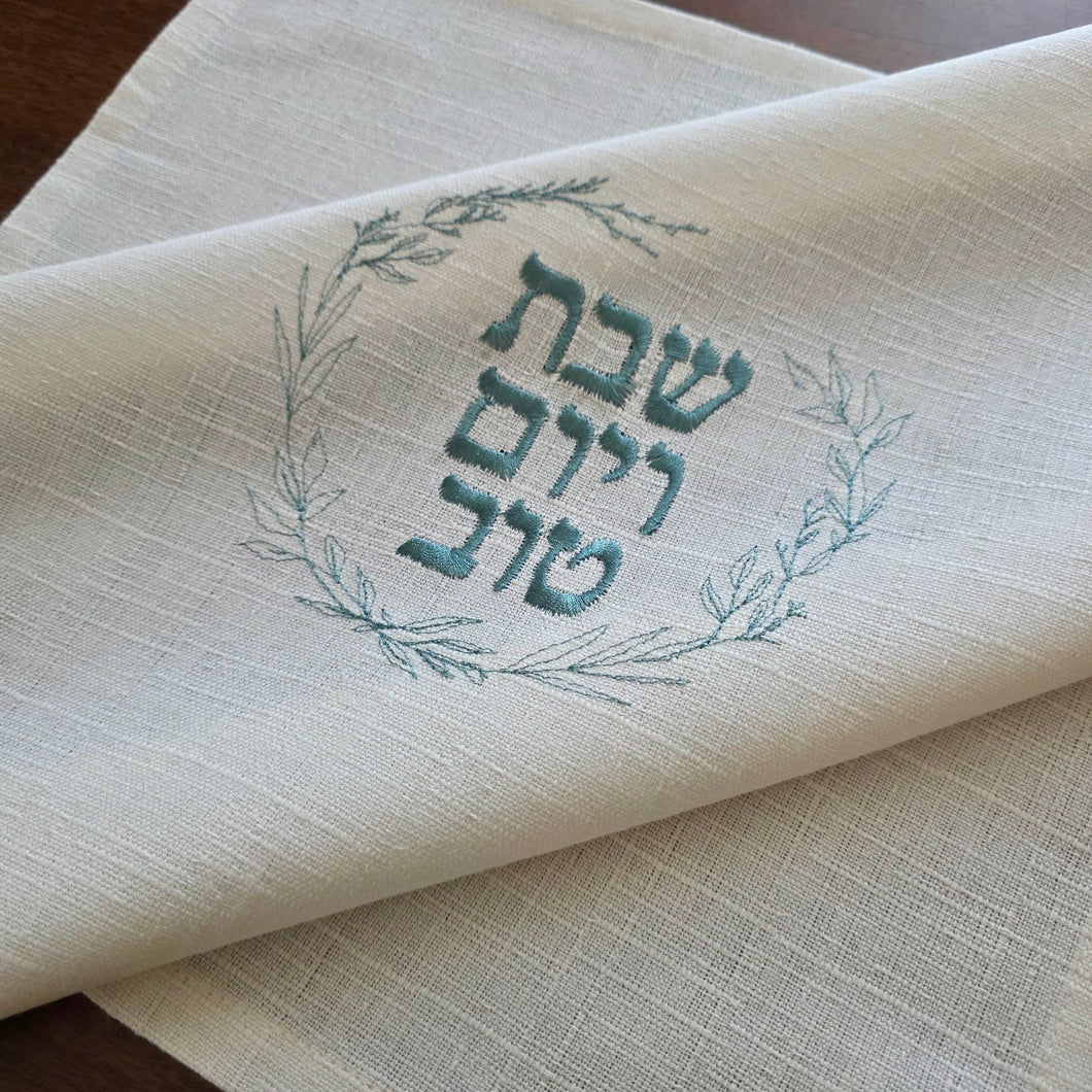 A woven cotton-blend challah cover featuring blue sage embroidered wreath with the message Shabbat v’Yom Tov in Hebrew lettering. Measures 18” x 18”.