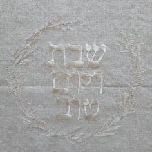 Closeup detail of Challah Cover embroidery in soft white stitching on a natural white woven fabric background with subtle silver flecks. Embroidery features a leafy wreath design surrounding the words Shabbat v’Yom Tov in Hebrew lettering.