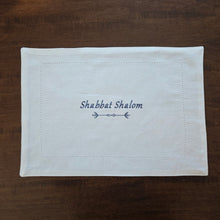 A white, woven, rectangular challah cover is shown. It features the words Shabbat Shalom embroidered horizontally across the center, above a geometric double-arrow in navy blue. There is an additional, white, keyhole-embroidery hem-stitch inset detail as well, bordering the challah cover.