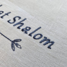 A closeup detail of the white, woven Shabbat Shalom Challah Cover embroidery. The portion of the message shown says Shalom over a geometric design.