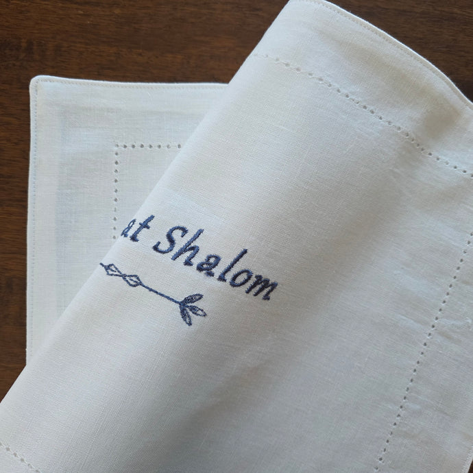 A white, woven cotton-blend challah cover features the words Shabbat Shalom embroidered in English letters above a simple geometric double-arrow design in navy blue.