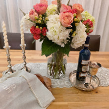 A Shabbat table set with a floral arrangement beside a silver wine tray and kiddush cup, a pair of silver candlesticks and a wooden bread board with a loaf of challah and a natural white challah cover embellished with pearl edging and the words Shabbat v’Yom Tov embroidered in the center in soft white thread, encircled by a leafy wreath design in matching thread.