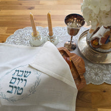A table set with white hydrangeas in a silver pitcher next to a silver kiddush cup and a pair of candlesticks on a sparkling table runner. A bread board bears a challah with a natural, soft-white cover, decorated with a leafy blue-green wreath around the Hebrew words Shabbat v’Yom Tov.