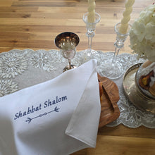 A Shabbat table is set with white hydrangeas in a silver pitcher on a sparkly table runner beside a pair of glass candlesticks and a silver kiddush cup, along with a wooden serving tray holding a loaf of bread covered by a white woven challah cover. The cover is embroidered with the words Shabbat Shalom in navy blue.