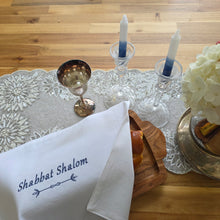 A Shabbat table is set with white hydrangeas in a silver pitcher on a sparkly table runner beside a pair of glass candlesticks and a silver kiddush cup, along with a wooden serving tray holding a loaf of bread covered by a white woven challah cover. The cover is embroidered with the words Shabbat Shalom in navy blue.