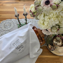 A Shabbat table set with a floral arrangement, a silver wine tray and kiddush cup and a pair of painted metal rose candlesticks beside a bread tray with a braided loaf covered by a white, woven Challah Cover with picot edging featuring center embroidery. Silver embroidery spells out Shabbat v’Yom Tov in Hebrew lettering encircled by a leafy wreath in the same silver color