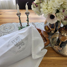 A Shabbat table set with a floral arrangement, a silver wine tray and kiddush cup and a pair of painted metal rose candlesticks beside a bread tray with a braided loaf covered by a white, woven Challah Cover with picot edging featuring center embroidery. Silver embroidery spells out Shabbat v’Yom Tov in Hebrew lettering encircled by a leafy wreath in the same silver color.