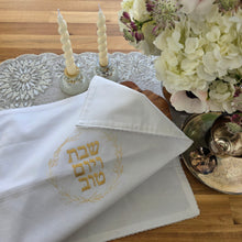 A Shabbat table is set with a floral arrangement, a silver wine tray and kiddush cup, a pair of glass candlesticks and a white, woven Challah Cover with picot edging featuring center embroidery. Gold embroidery spells out Shabbat v’Yom Tov in Hebrew lettering encircled by a leafy wreath in the same gold color.