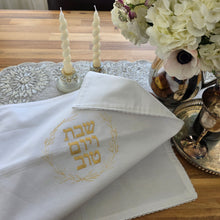 A Shabbat table is set with a floral arrangement, a silver wine tray and kiddush cup, a pair of glass candlesticks and a white, woven Challah Cover with picot edging featuring center embroidery. Gold embroidery spells out Shabbat v’Yom Tov in Hebrew lettering encircled by a leafy wreath in the same gold color.