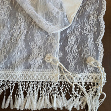 White lace prayer shawl with intricate floral patterns, a decorative Venetian lace trim, and hanging tzitzit fringes, laid flat on a dark brown surface.