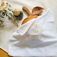 White, woven cotton-blend challah cover with petite pom-pom fringe and gold embroidered Shanah Tova text in Hebrew encircled by a leafy gold embroidered wreath design. Shown on an apple-shaped wooden challah board alongside a glass honey server.