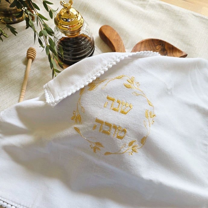 White, woven cotton-blend challah cover with petite pom-pom fringe and gold embroidered Shanah Tova text in Hebrew encircled by a leafy gold embroidered wreath design. Shown on an apple-shaped wooden challah board alongside a glass honey server.