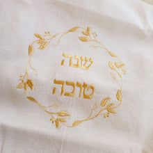 White, woven cotton-blend challah cover with gold embroidered Shanah Tova text in Hebrew encircled by a leafy gold embroidered wreath design. 