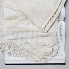 A handmade lace tallit in soft eggnog tones lays on a book of Torah text.